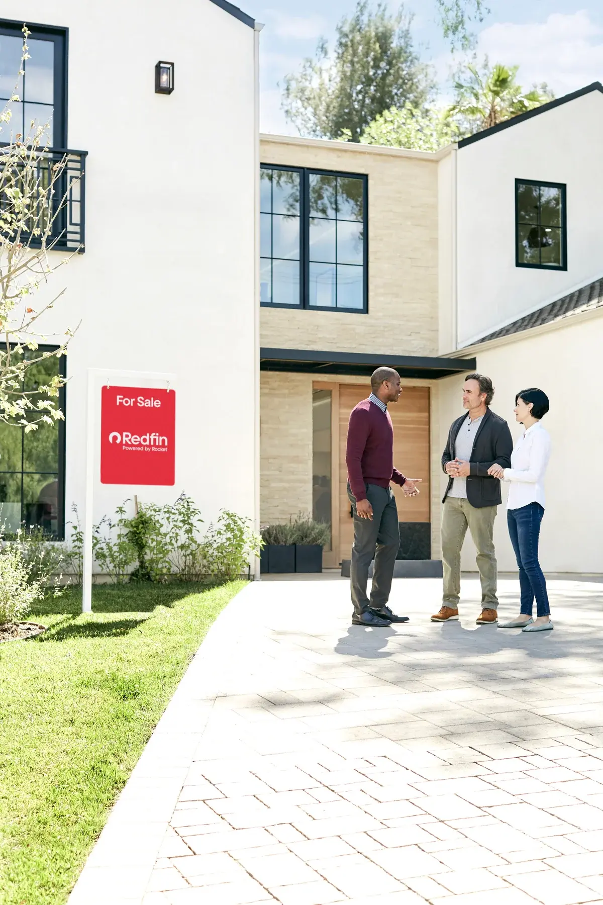 Three people outside a modern home beside a Redfin for-sale sign